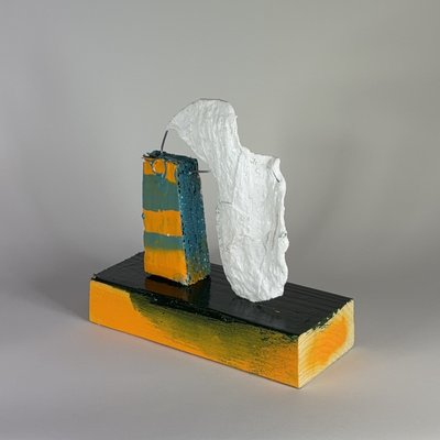 Angled view of a textured white face suspended by steel wires beside a teal and yellow upright form, on a black-topped yellow base.